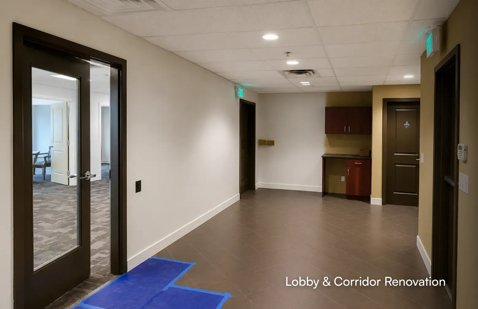 Renovated commercial office lobby and corridor featuring modern brown floor tiles, dark wood doors, and a built-in cabinetry station, completed by PCC - Pacific Coast Contracting.